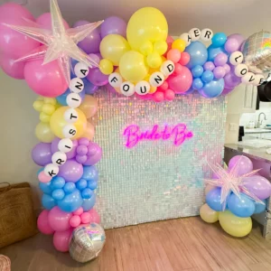 In Her Bach Era bachelorette backdrop with iridescent shimmer wall, pastel rainbow balloon garland, friendship bracelet balloon banner, and Bride to Be neon sign