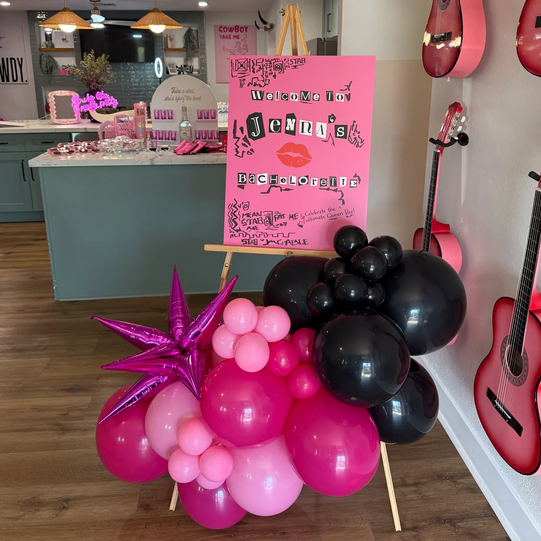 Mean Girls bachelorette welcome sign with pink Burn Book style design and black balloon garland on easel